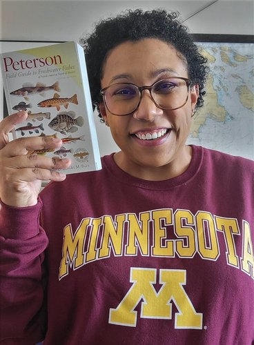 smiling woman wearing glasses and a U of M top holding a book on fishes